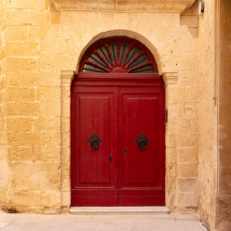 Red arched double door with black ironwork in a limestone wall in Mdina, Malta.