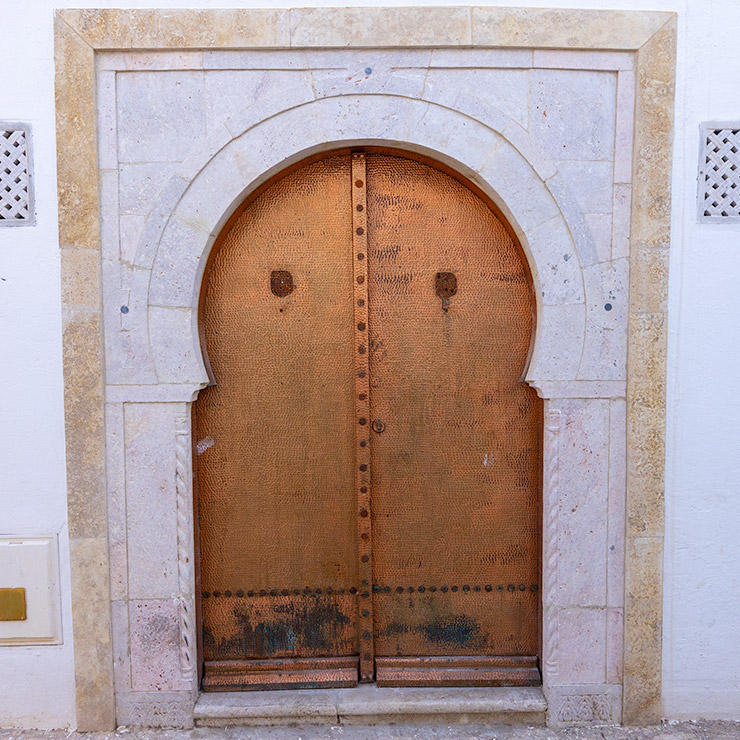 Bronze arched door with a hammered texture set in a pale stone frame, located in the Medina of Tunis.