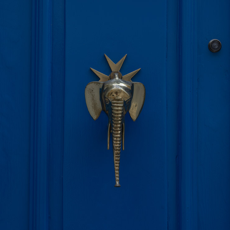 Polished brass elephant-shaped doorknob mounted on a vivid blue door in Birgu, Malta, featuring a long downward-curving trunk and stylized sunburst or crown motif above its head.