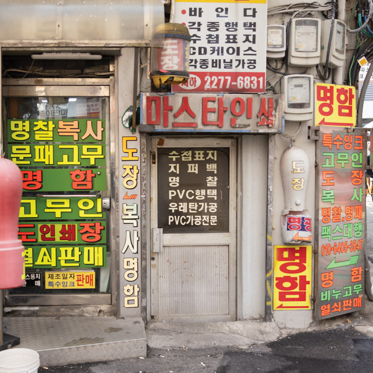 A narrow, weathered door in an Euljiro alley in Seoul, surrounded by colorful, densely packed Korean signage advertising printing, name tag, and seal services. The small storefront is cluttered with layered posters and old utility meters, capturing the gritty, industrious charm of the neighborhood.