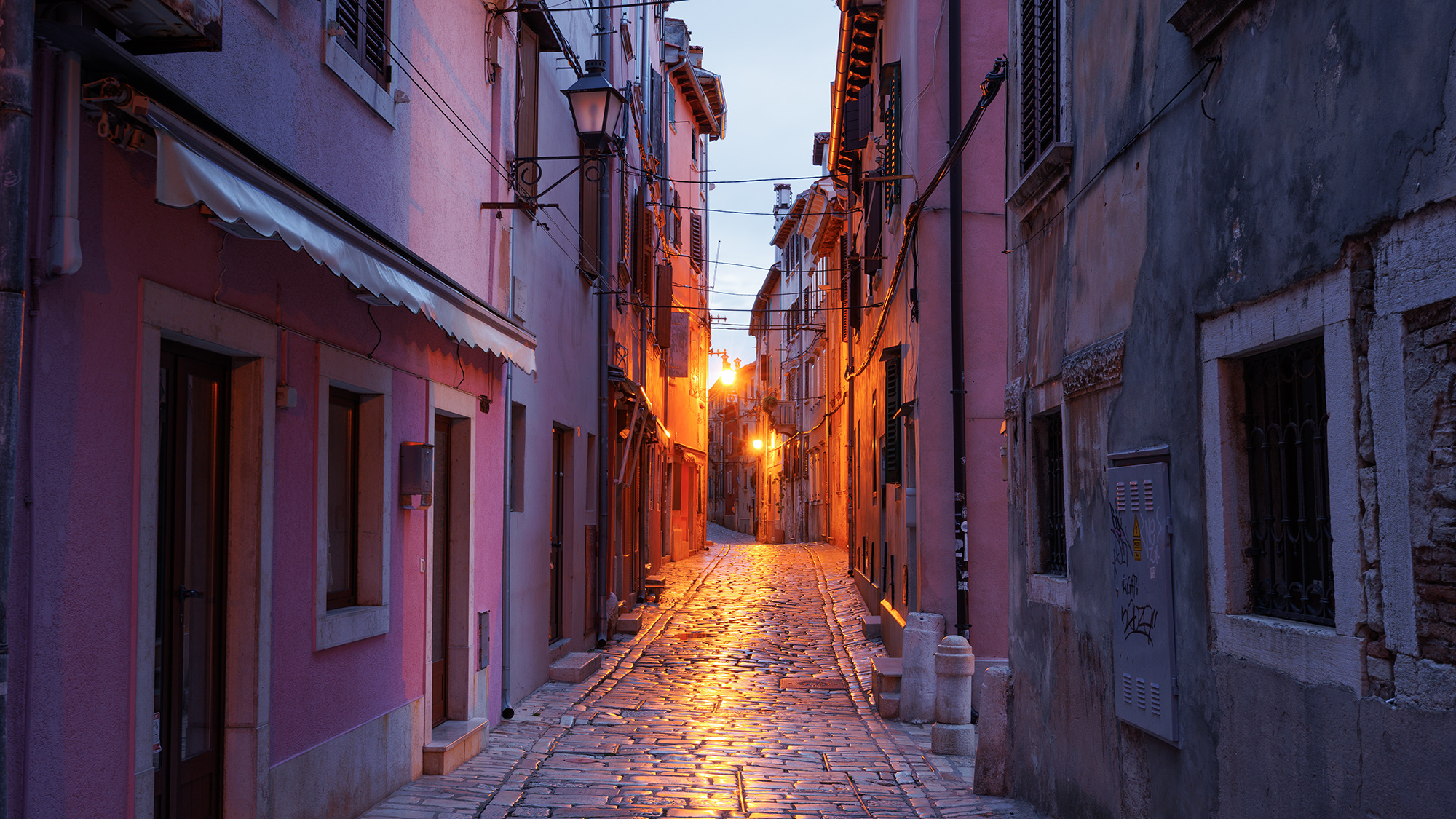 Narrow dusk alley in Rovinj, Croatia—warm stone walls, glowing windows and lantern light, polished cobbles tapering toward the sea—an invitation to wander and discover Europe.