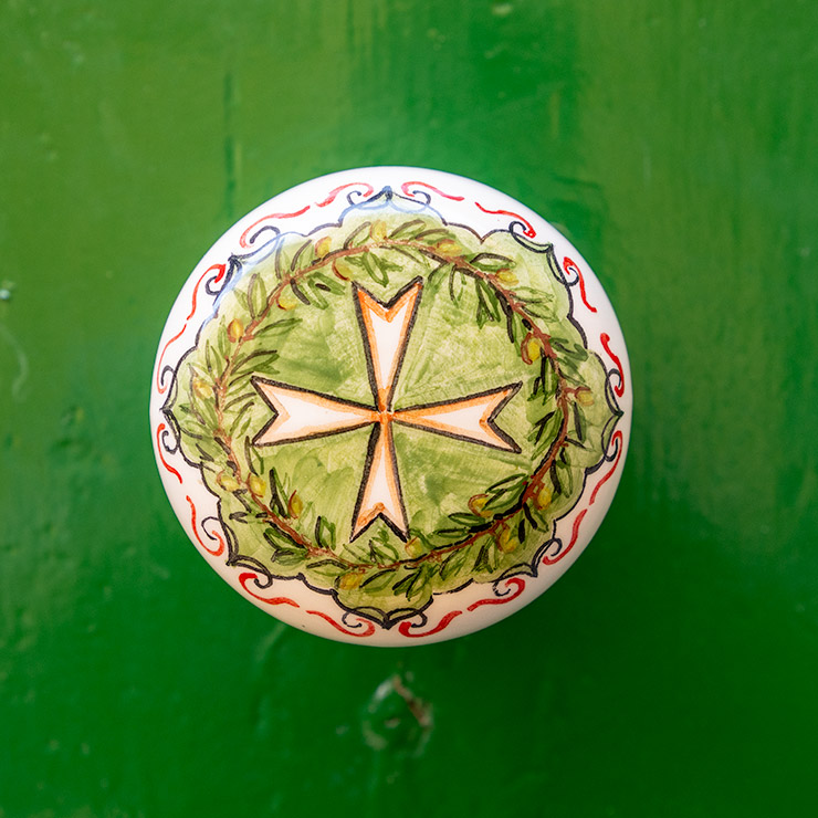 Hand-painted ceramic door knob on a green wooden door in Birgu, Malta, featuring a traditional eight-pointed Maltese Cross encircled by a laurel wreath and bordered with red and blue scrollwork.
