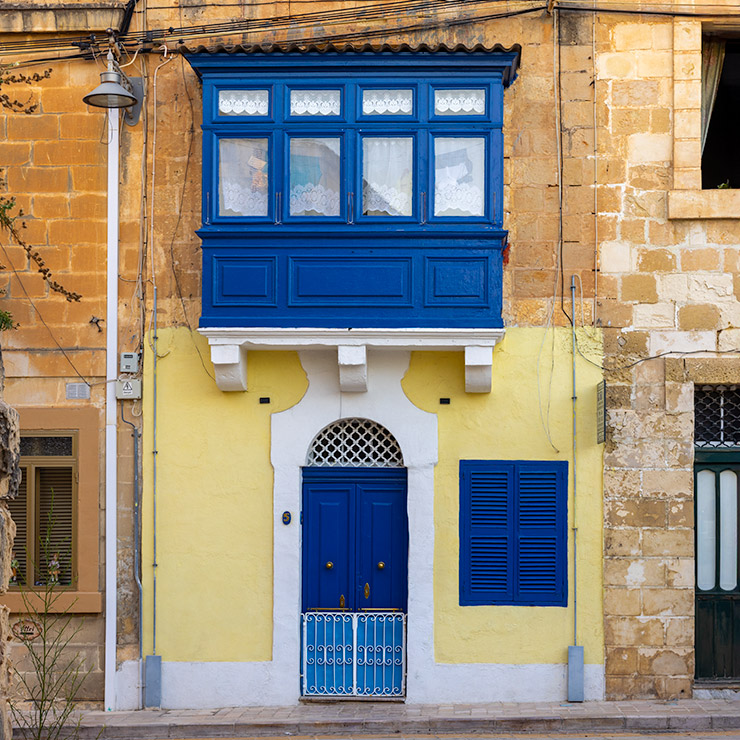 Bold blue Maltese door and matching shuttered window set into a yellow-painted stone façade in Mdina, Malta, topped by a traditional enclosed wooden balcony with lace-curtained windows and white corbels.