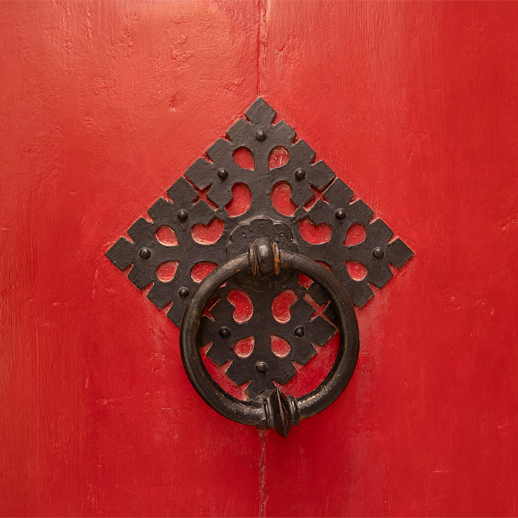 Ornate black iron ring door knocker on a bold red wooden door in Mdina, Malta, mounted on a decorative diamond-shaped iron backplate with intricate cutouts and small raised rivets.
