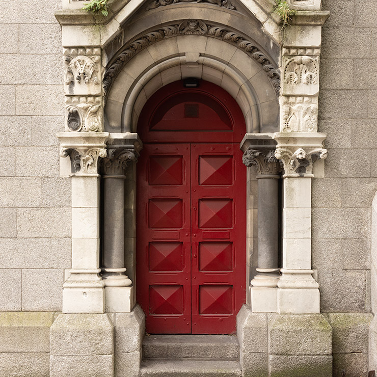 Red double doors set within a Romanesque stone archway with carved columns and decorative capitals at St. Andrew's Church in Dublin, Ireland.