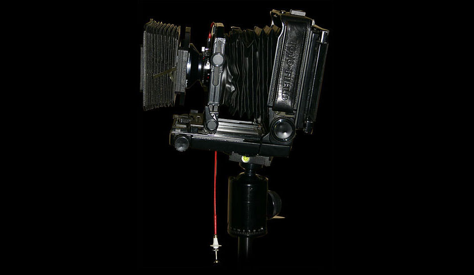 Side view of a Toyo 45AX large format field camera mounted on a tripod, with bellows extended and a red shutter release cable hanging below. The camera is set against a black background.