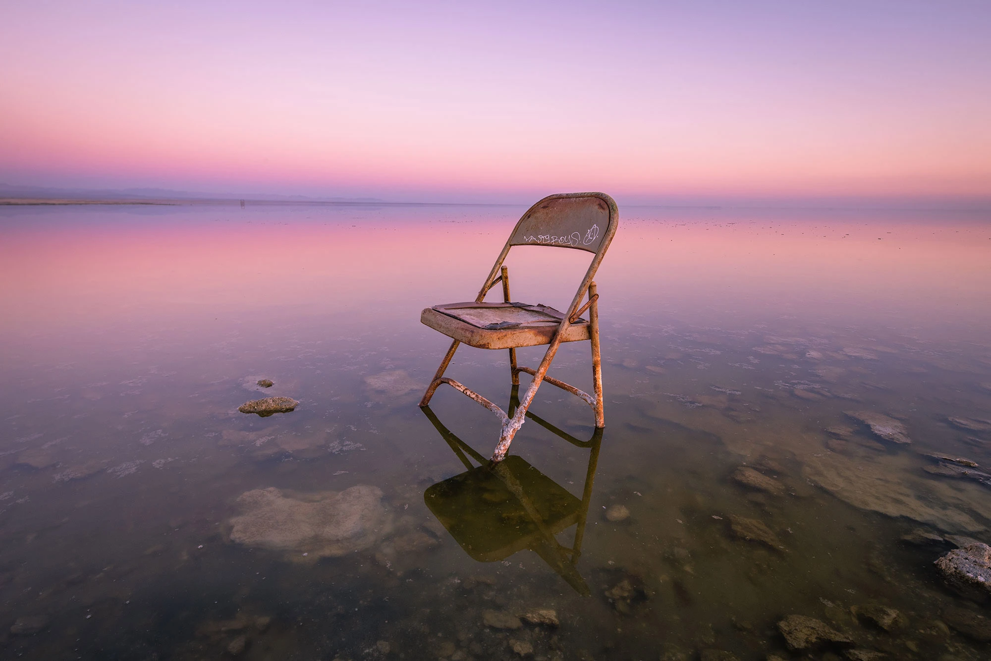 A lone chair sinking into the sand on the desolate shoreline of Bombay Beach, California, its rusted frame and peeling paint symbolizing abandonment and the eerie beauty of the Salton Sea's decline.