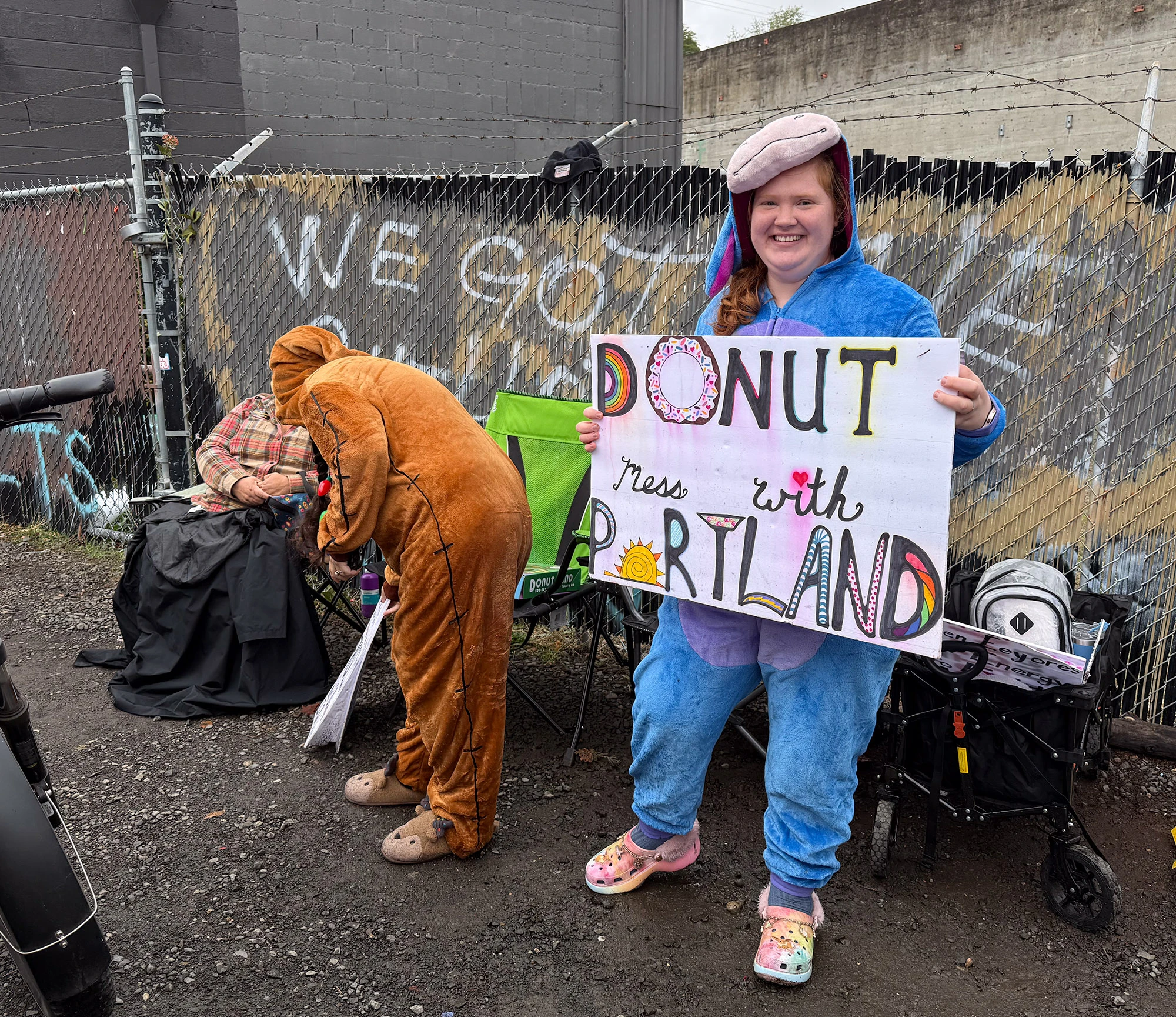 A woman in a blue onesie and colorful Crocs stands smiling in front of Portland's ICE building, holding a handmade sign that reads 'DONUT mess with PORTLAND' — a mix of humor and defiance that captures the city's creative protest spirit.