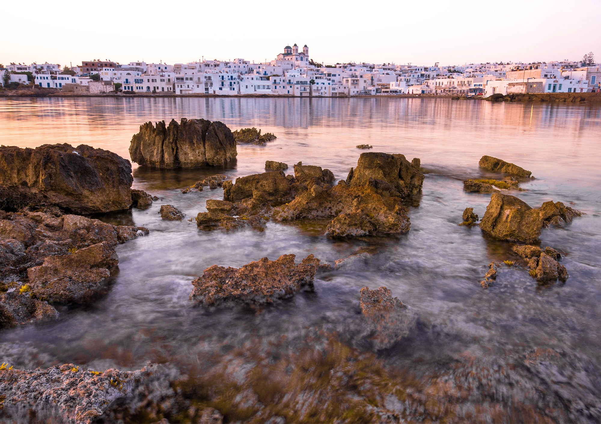 Fishing boats float in the serene harbor of Naoussa, Paros, their hulls casting colorful reflections on the still Aegean water. The Cycladic architecture glows in early morning light, with whitewashed walls and blue accents framing the peaceful scene.