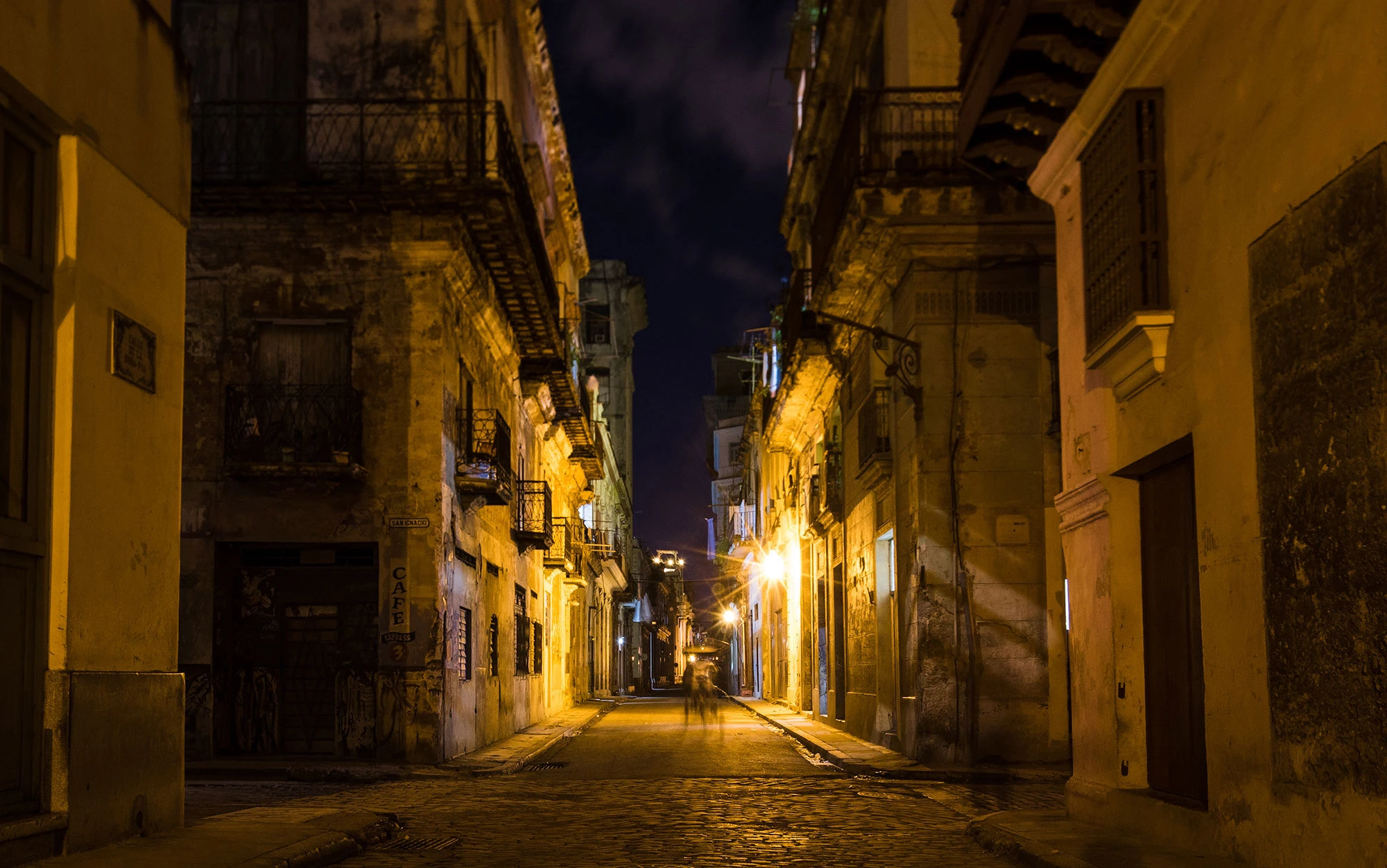 Night in Habana Vieja: a narrow cobblestone street washed in amber lamplight, weathered colonial facades and wrought-iron balconies receding to a vanishing point, with long-exposure silhouettes of late-night walkers.