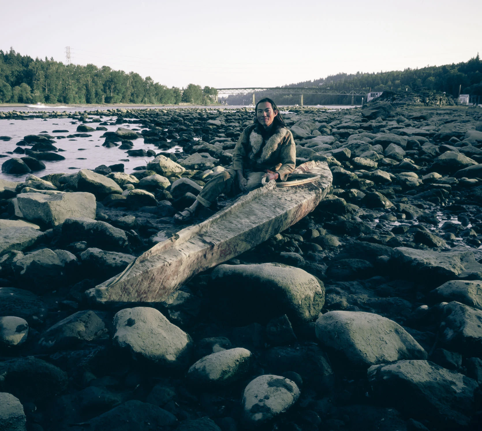 Primitivist Kiliii sitting on a hand-carved dugout canoe among river rocks on the Willamette River shoreline