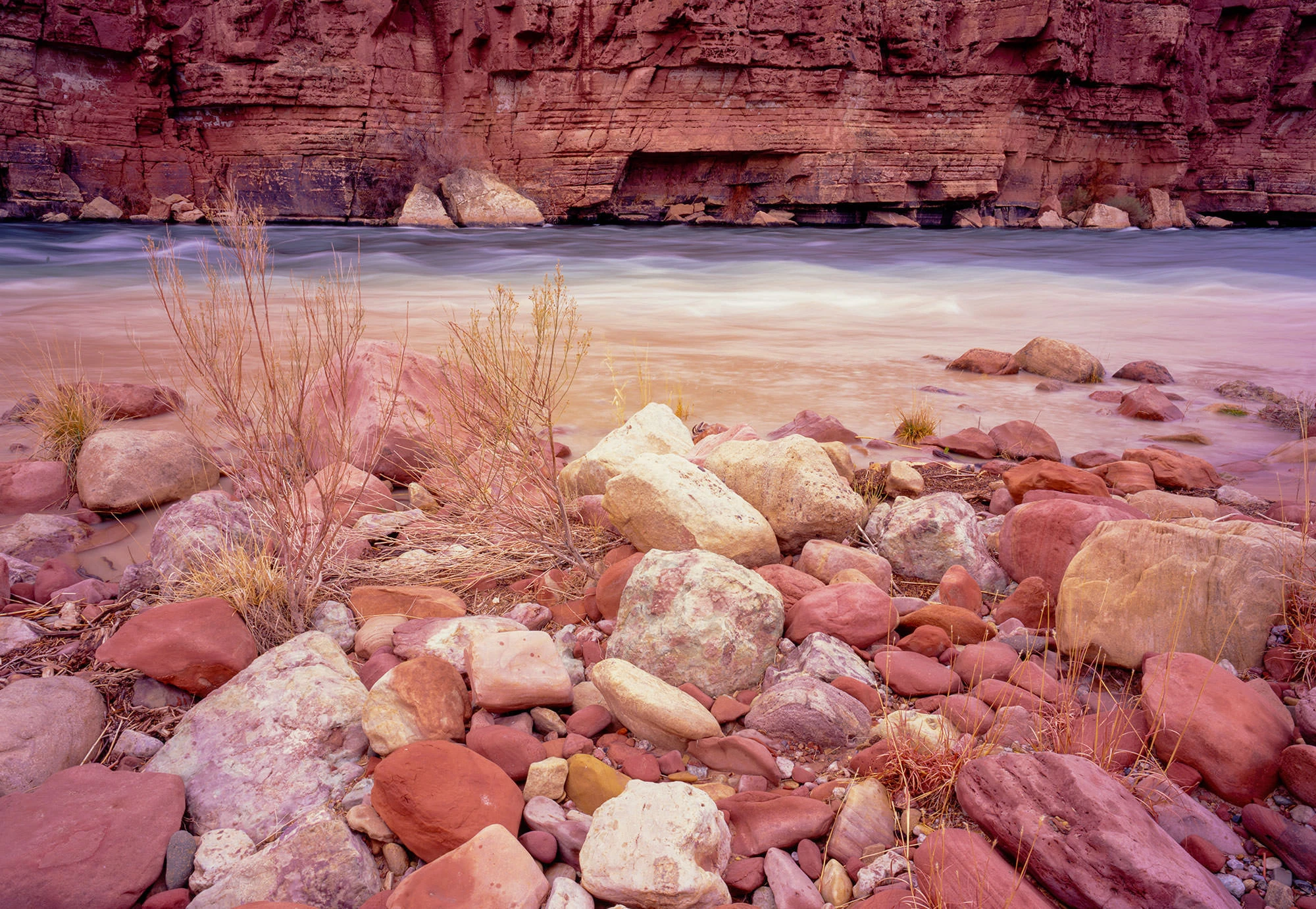 Soft-focus long exposure of the Colorado River at Lees Ferry, the water appearing misty and blurred as it flows beneath high canyon walls. The scene is framed by red sandstone and sparse desert vegetation, with the river leading the eye downstream into the Vermilion Cliffs wilderness.