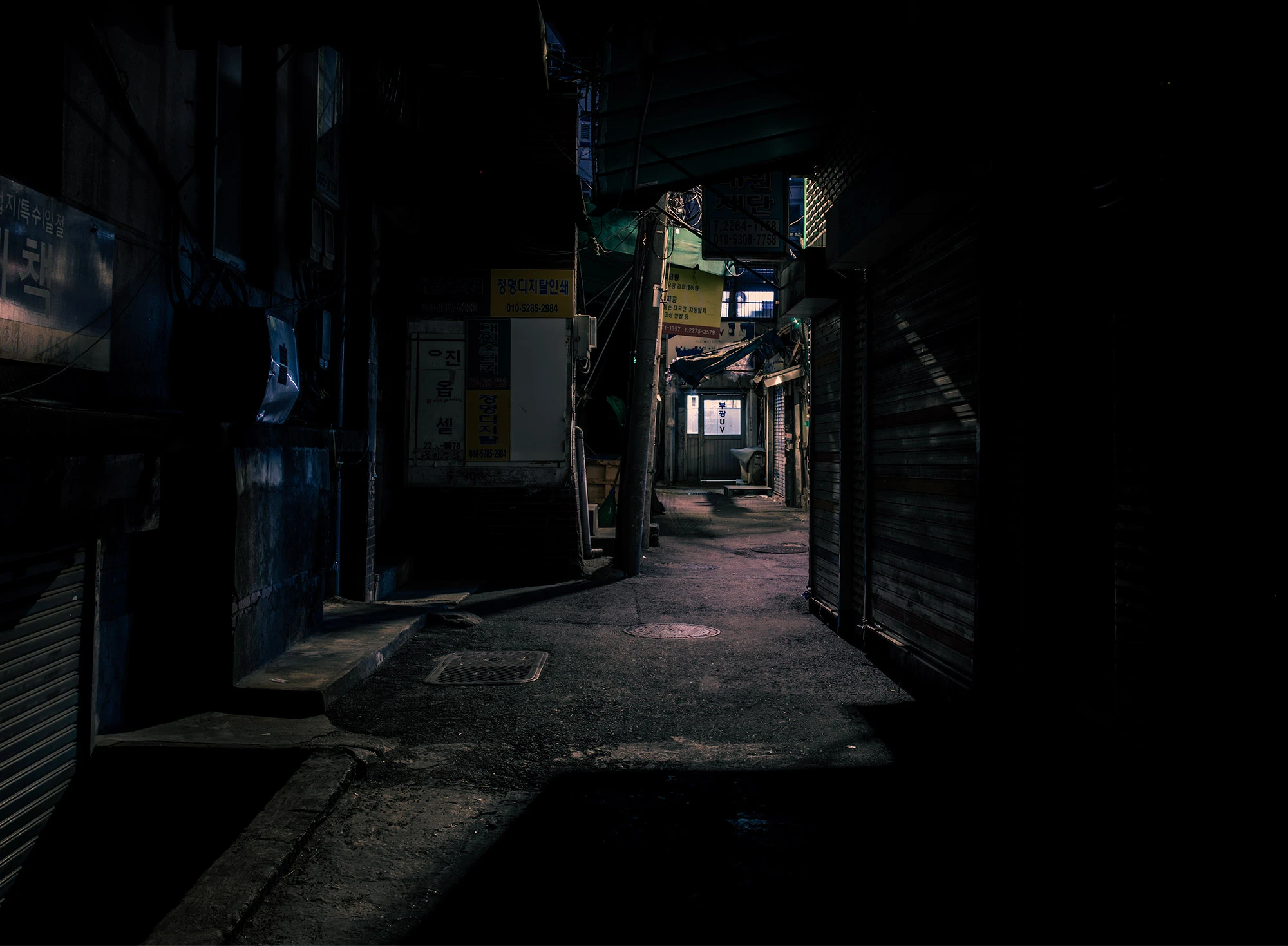 A dimly lit alley in Seoul's Euljiro neighborhood winds between tightly packed buildings, with most storefronts shuttered and dark. A faint bluish glow from a distant window and scattered neon signs hints at life beyond the shadows, casting moody reflections on the cracked pavement.