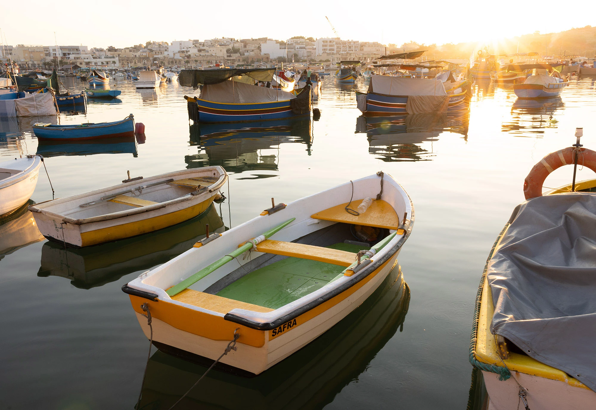 Rowboats and traditional luzzu fishing boats float in the golden light of sunrise at Marsaxlokk Harbor, Malta, their reflections rippling across the still water.