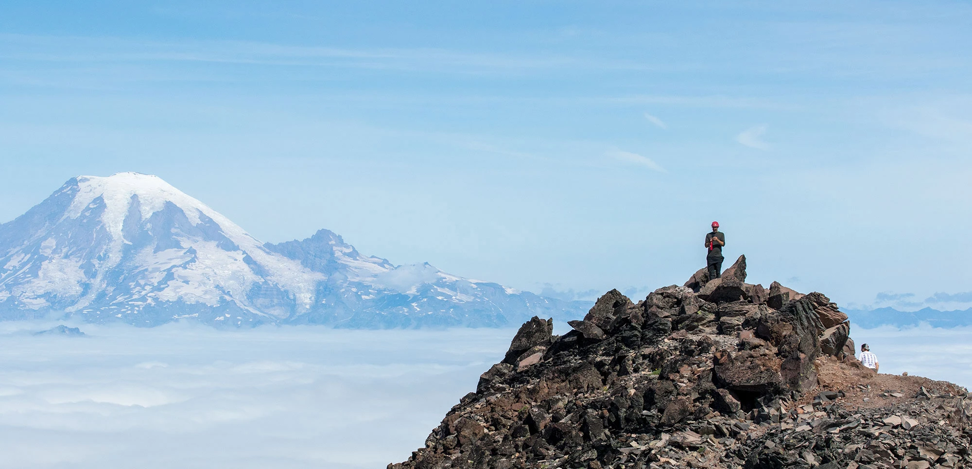Backpacking companions Eric and Tim pause on the rocky summit ridge of Old Snowy Peak in Washington's Goat Rocks Wilderness, looking out over a sea of clouds toward the massive snow-covered dome of Mount Rainier. The jagged volcanic rock foreground contrasts with the sweeping alpine view, capturing both the scale of the Cascade Range and the exhilaration of reaching the high point on a backcountry trek.