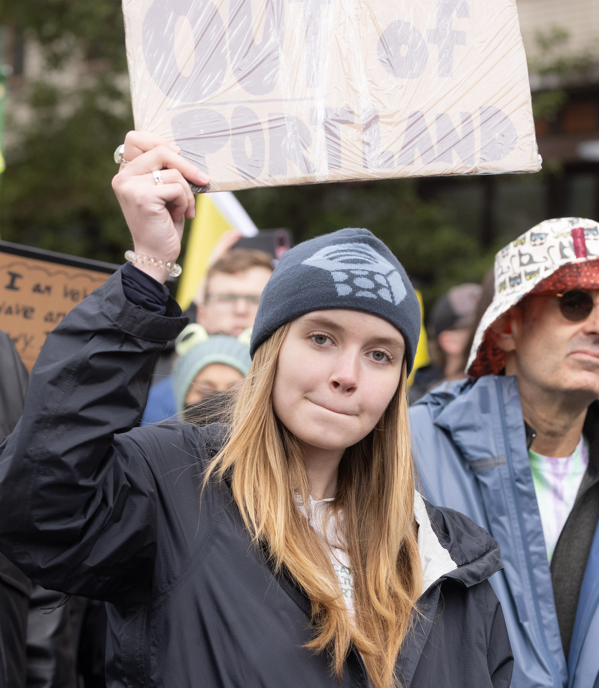At a Portland anti-ICE march, a young protester in a beanie raises a hand-made sign reading 'OUT OF PORTLAND' amid a packed crowd.