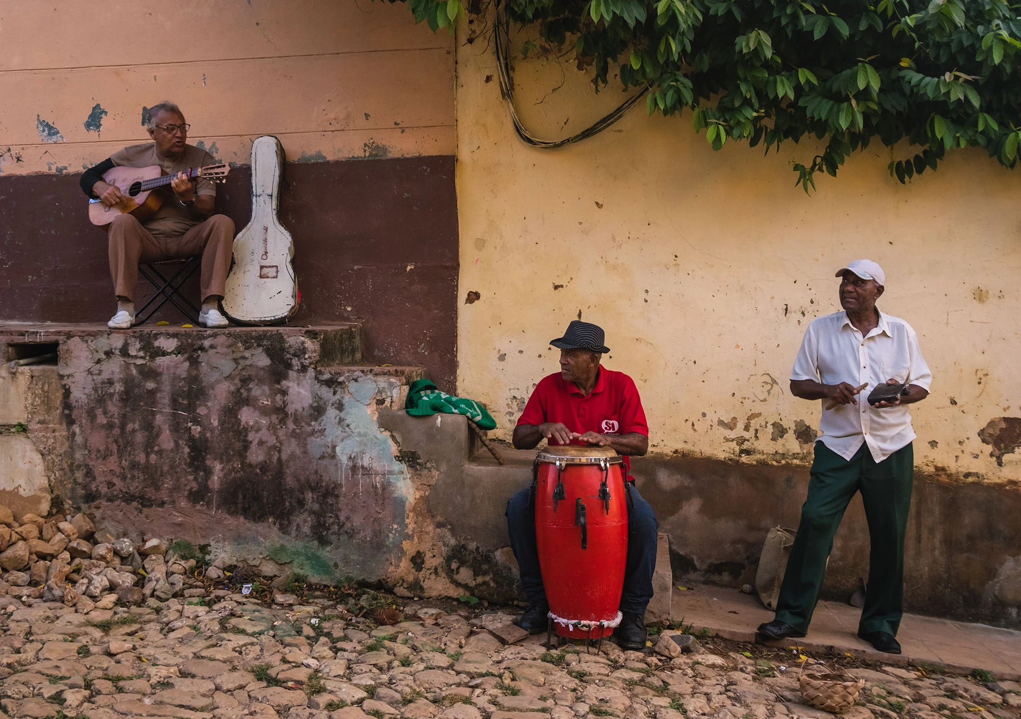 Three elderly Cuban musicians perform on a cobblestone street in Trinidad, Sancti Spíritus Province. One man plays a small guitar beside a weathered wall, another in a red shirt beats a tall red conga drum, and a third keeps rhythm on a metal guiro. Faded plaster, leafy vines, and the soft afternoon light add texture to this intimate glimpse of Cuba's street music culture.
