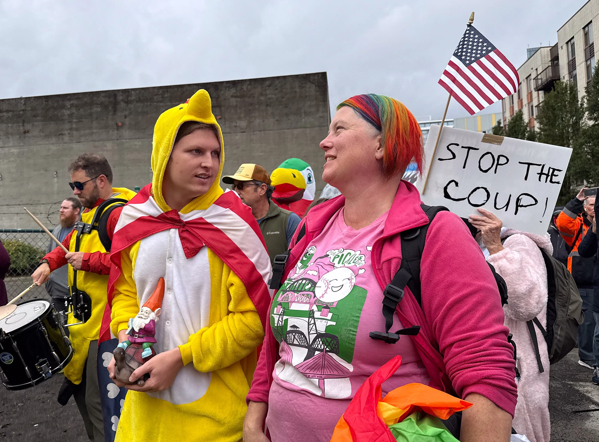 During Portland's ICE and National Guard protests, a man dressed in a yellow chicken costume — one of the first to appear in costume at these demonstrations — stands beside a woman with rainbow-colored hair and others carrying signs reading 'STOP THE COUP!' beneath a gray sky.