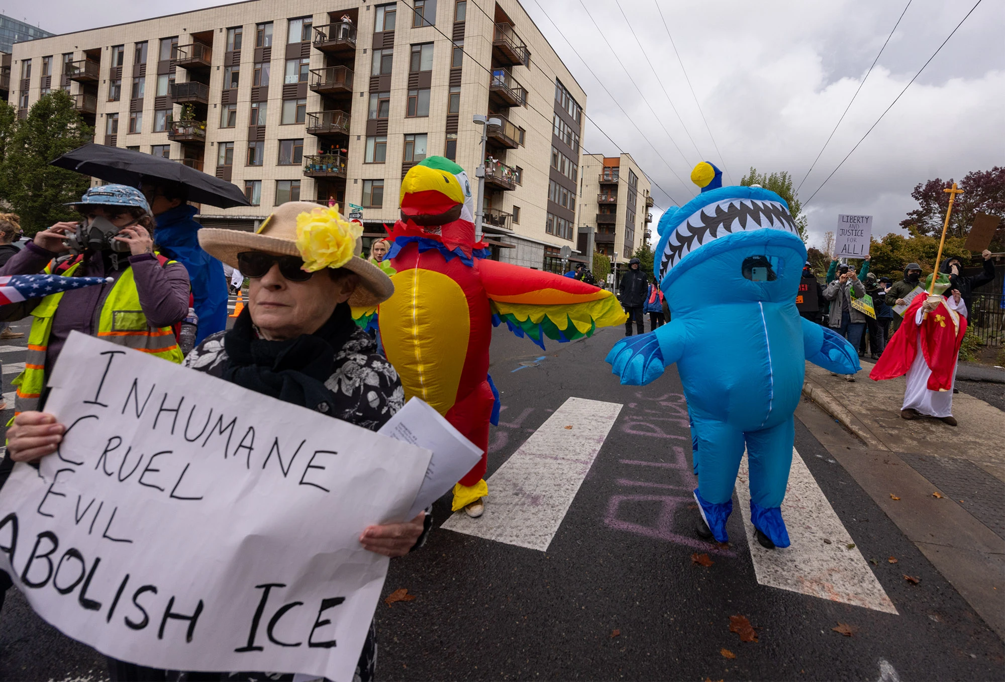 Protesters march through Portland's streets near the ICE building, combining humor and conviction. A woman in sunglasses holds a sign reading 'INHUMANE CRUEL EVIL ABOLISH ICE' beside people in inflatable shark and parrot costumes under a gray autumn sky.
