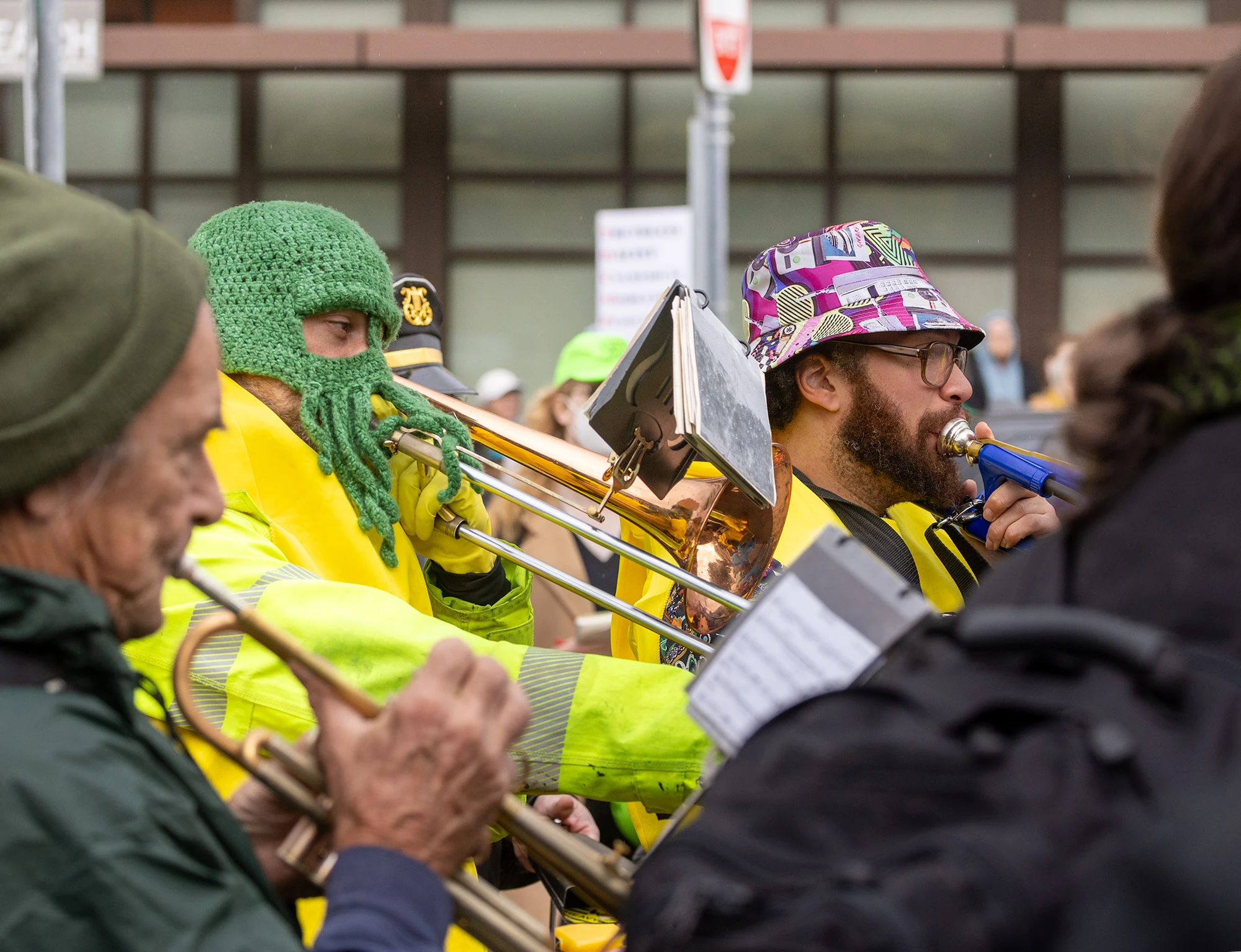 A protest band performs during Portland's anti-ICE demonstrations. A man in a green squid hat plays trombone beside another musician in a patterned bucket hat, bringing rhythm and humor to the march.