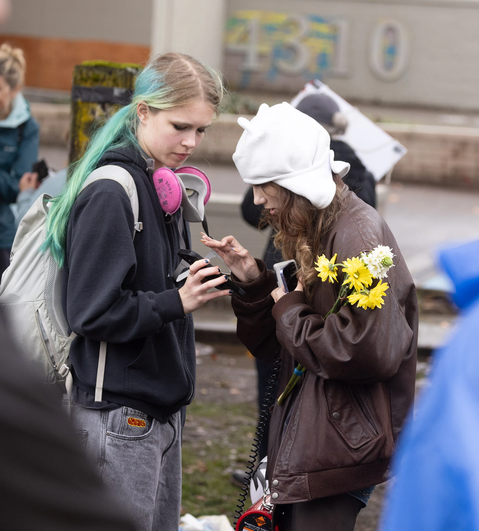 Two Portland protesters stand together outside the ICE building. One wears a hoodie and pink respirator, while the other, holding yellow flowers and a megaphone cord, leans in to share something on a phone amid the ongoing demonstration.