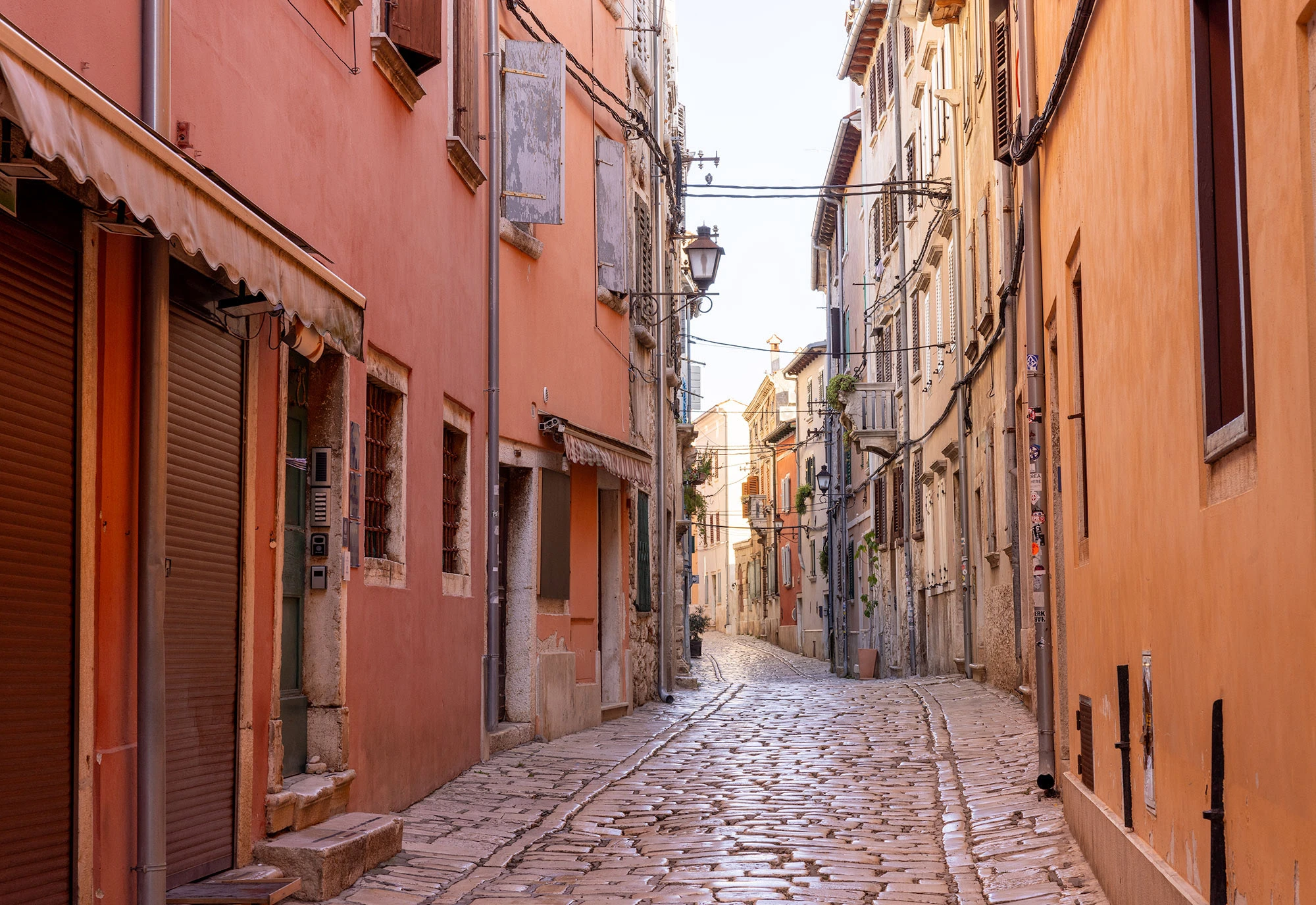 A predawn Rovinj alley with warm yellow-orange lighting, stone walls, and a blue-gray sky, creating a cozy, quiet atmosphere.