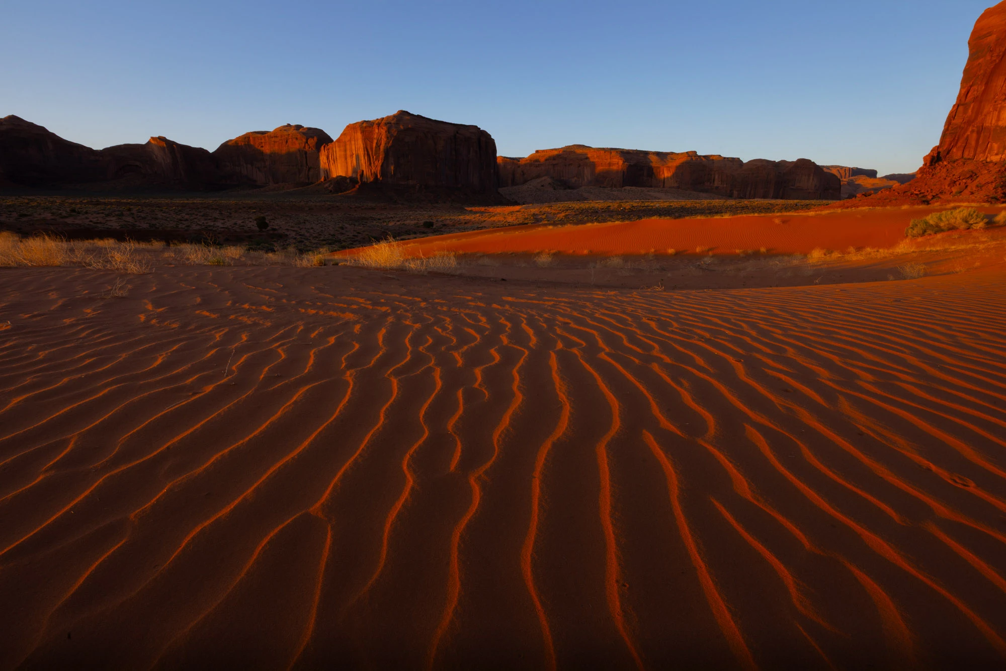 Rippling red sand dunes at Sand Springs in Monument Valley, with towering sandstone monoliths in the distance where Ancestral Puebloans once lived.
