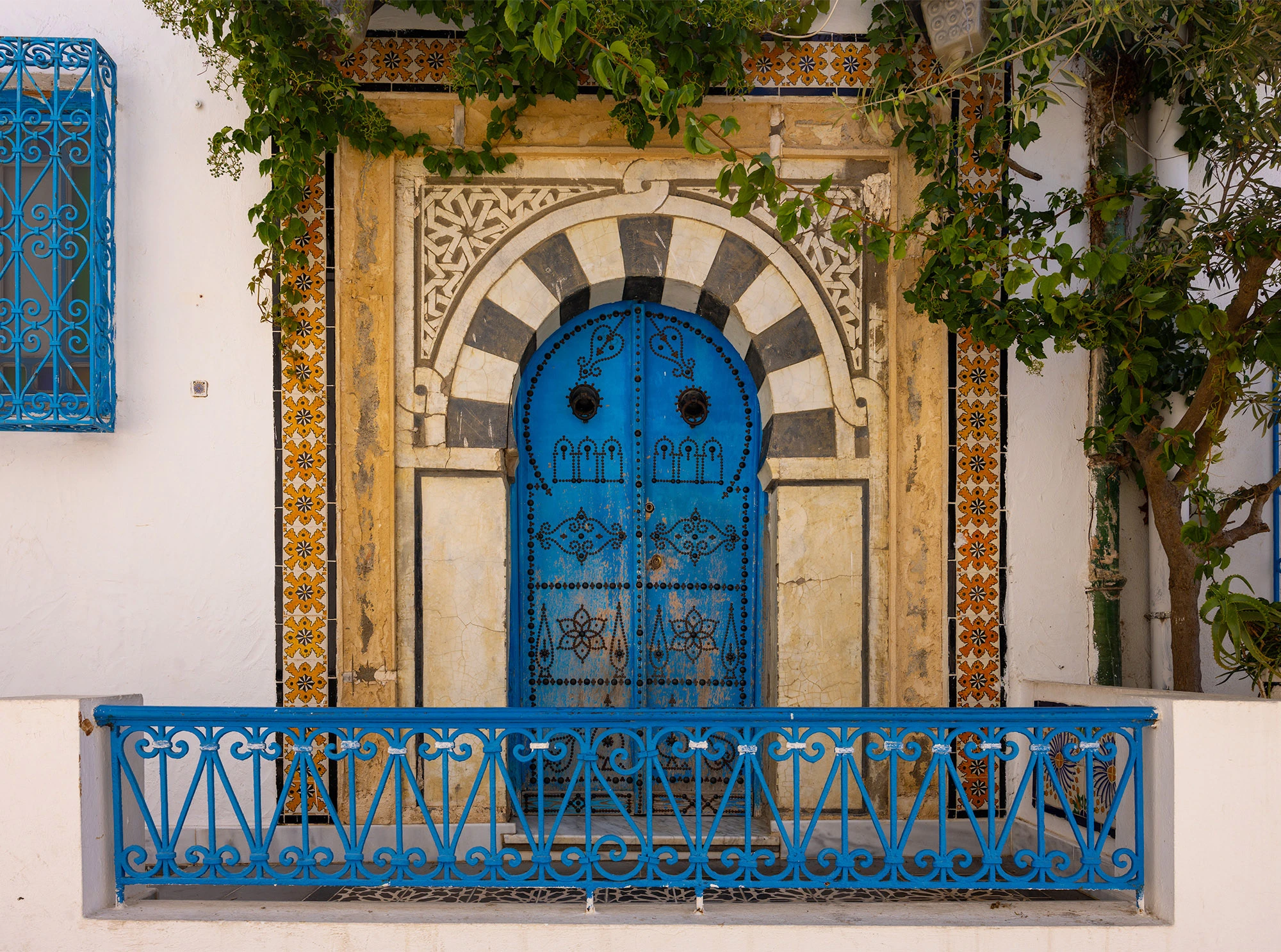 Traditional blue door in Sidi Bou Said framed by an arched limestone and marble doorway, patterned tiles, and cascading vines. The intricate ironwork and symmetry reflect the village's Andalusian-Maghrebi heritage and its enduring balance between ornament, function, and light.