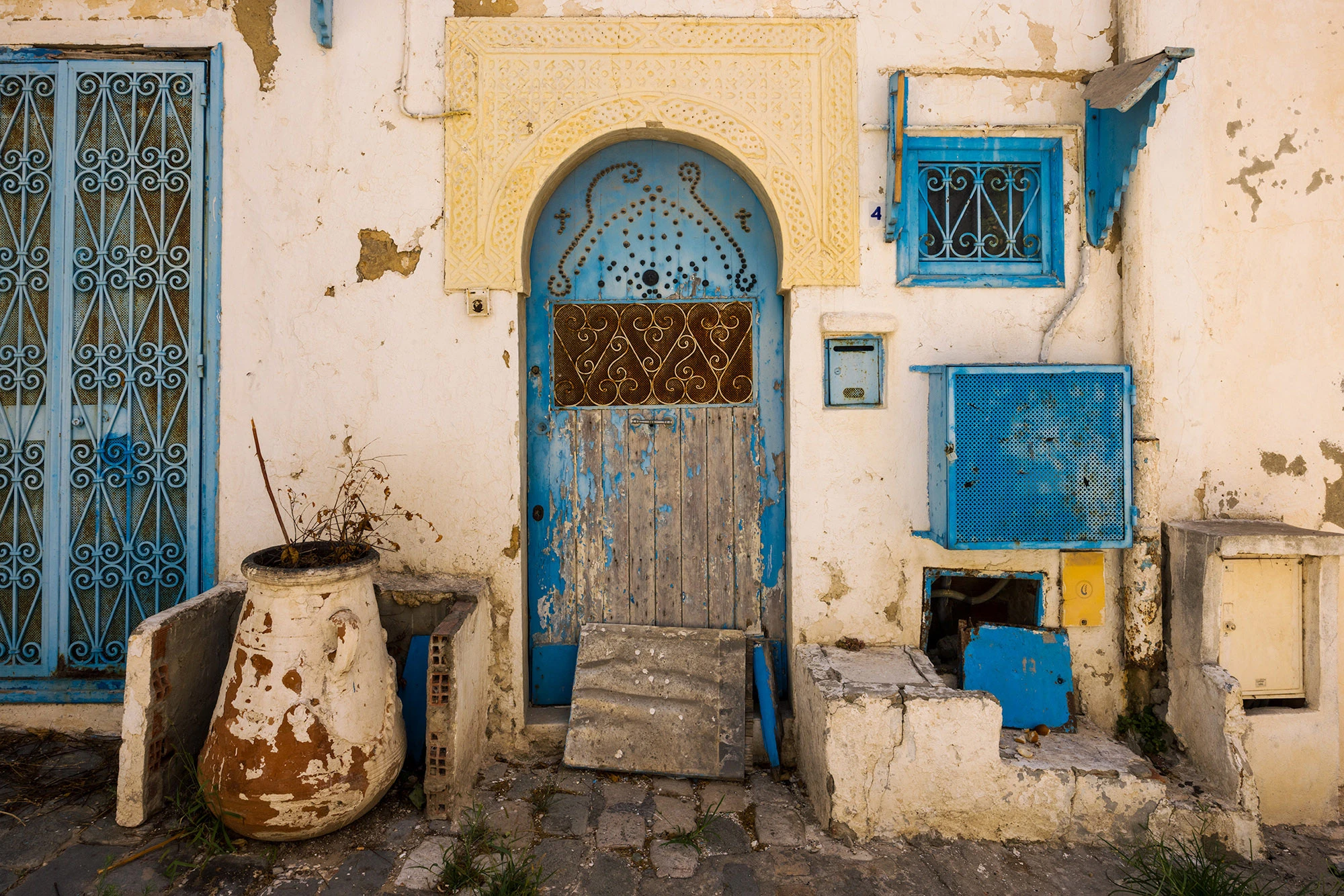 A weathered doorway in Sidi Bou Said, Tunisia, with peeling blue paint, carved plasterwork, and mismatched utilities framed by sun-faded walls. Signs of daily life—patched masonry, an old urn, and improvised repairs—reveal the lived-in texture that gives the village its enduring beauty and authenticity.