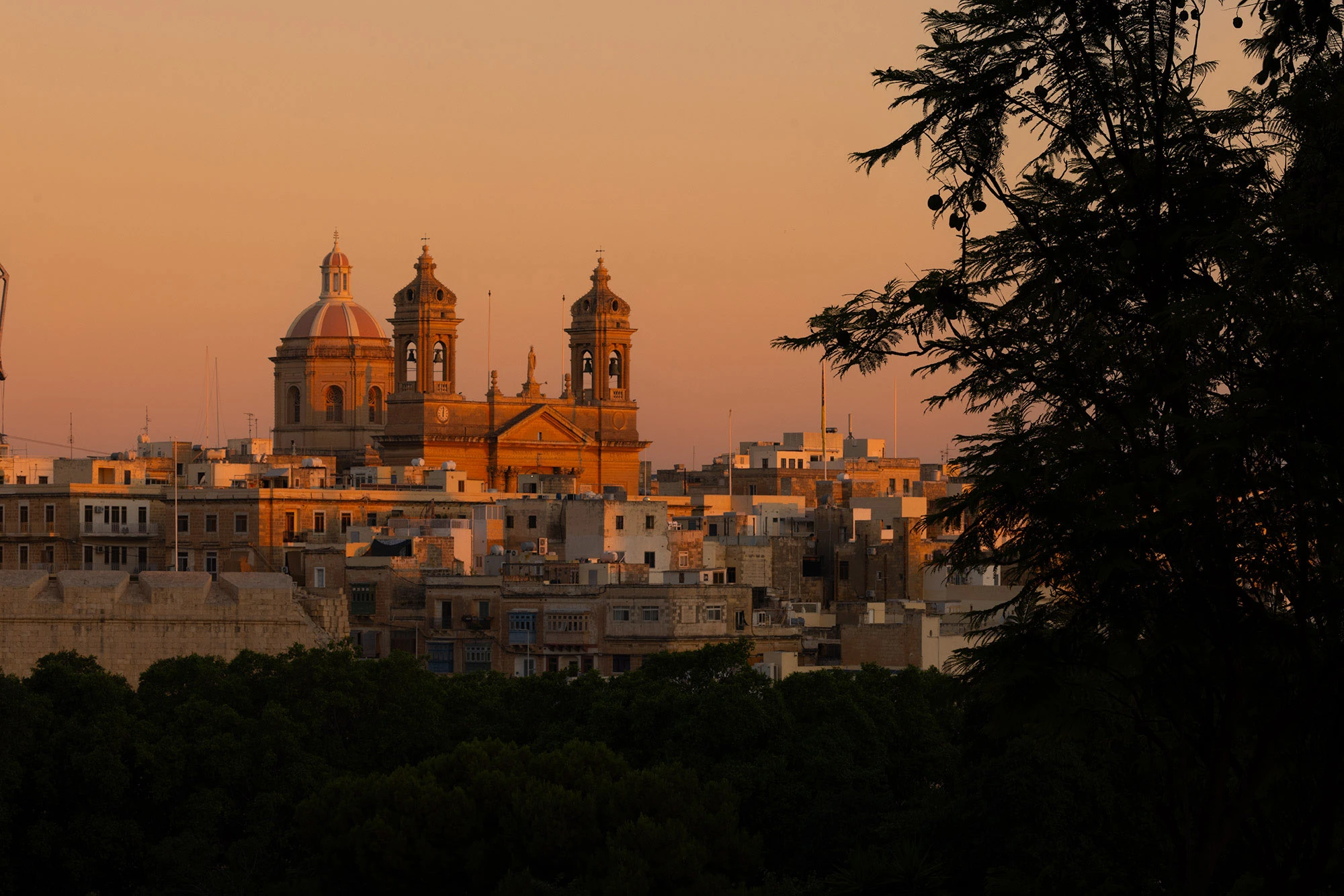 The skyline of Birgu, Malta, at sunset, with the dome and twin bell towers of St. Lawrence's Church glowing in golden light. The foreground is framed by dark trees in shadow, while limestone houses with flat roofs and painted balconies cluster tightly below the church. Once the headquarters of the Knights of St. John during the Great Siege, Birgu's silhouette still carries the weight of its history against the warm Mediterranean sky.