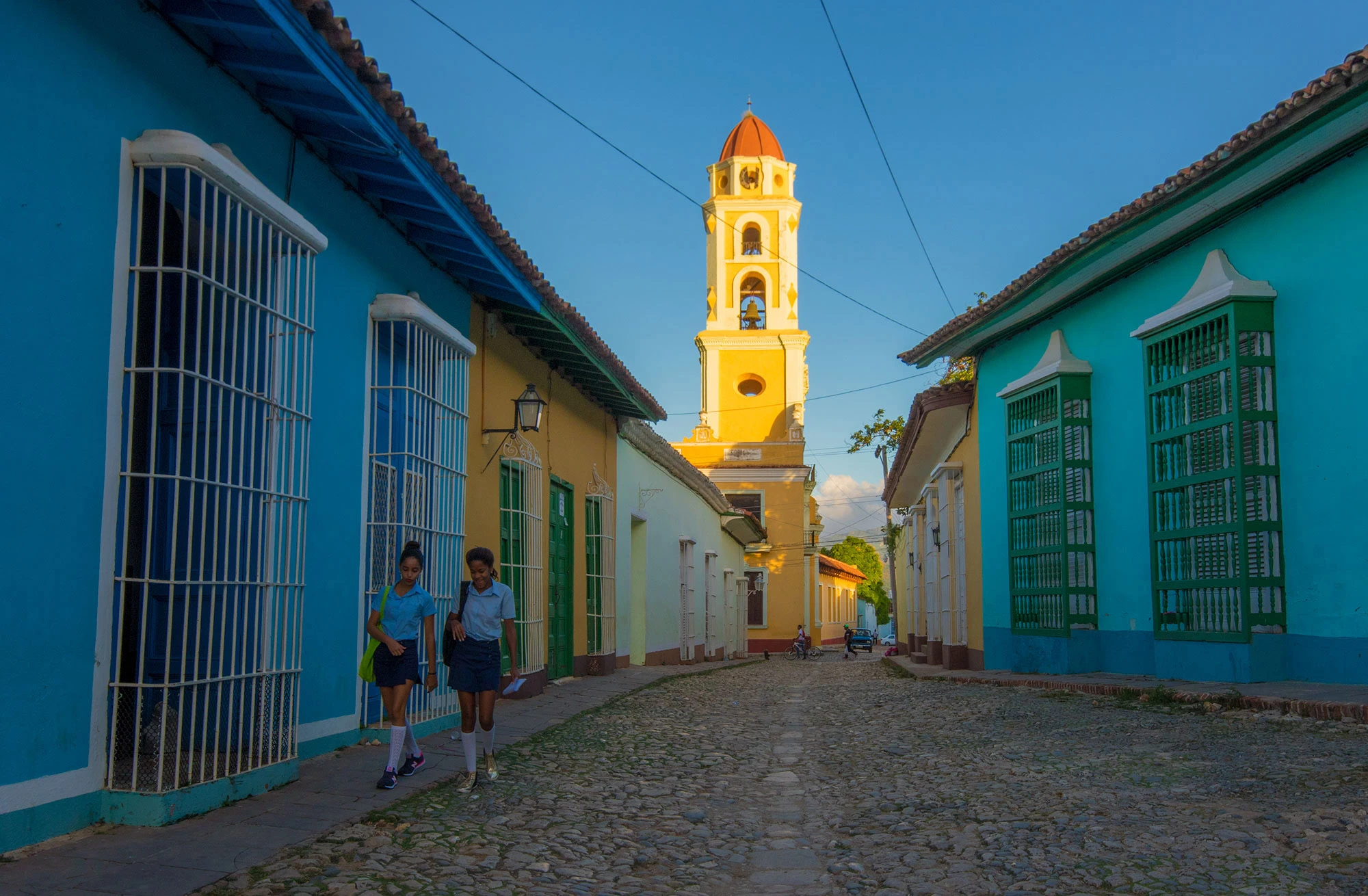 Two Cuban schoolgirls in blue uniforms walk down a cobblestone street in Trinidad, Sancti Spíritus Province, Cuba, passing brightly painted colonial houses. Behind them rises the yellow bell tower of the Convento de San Francisco de Asís, glowing in morning light that casts vivid contrasts of turquoise, ochre, and shadow across the historic town.