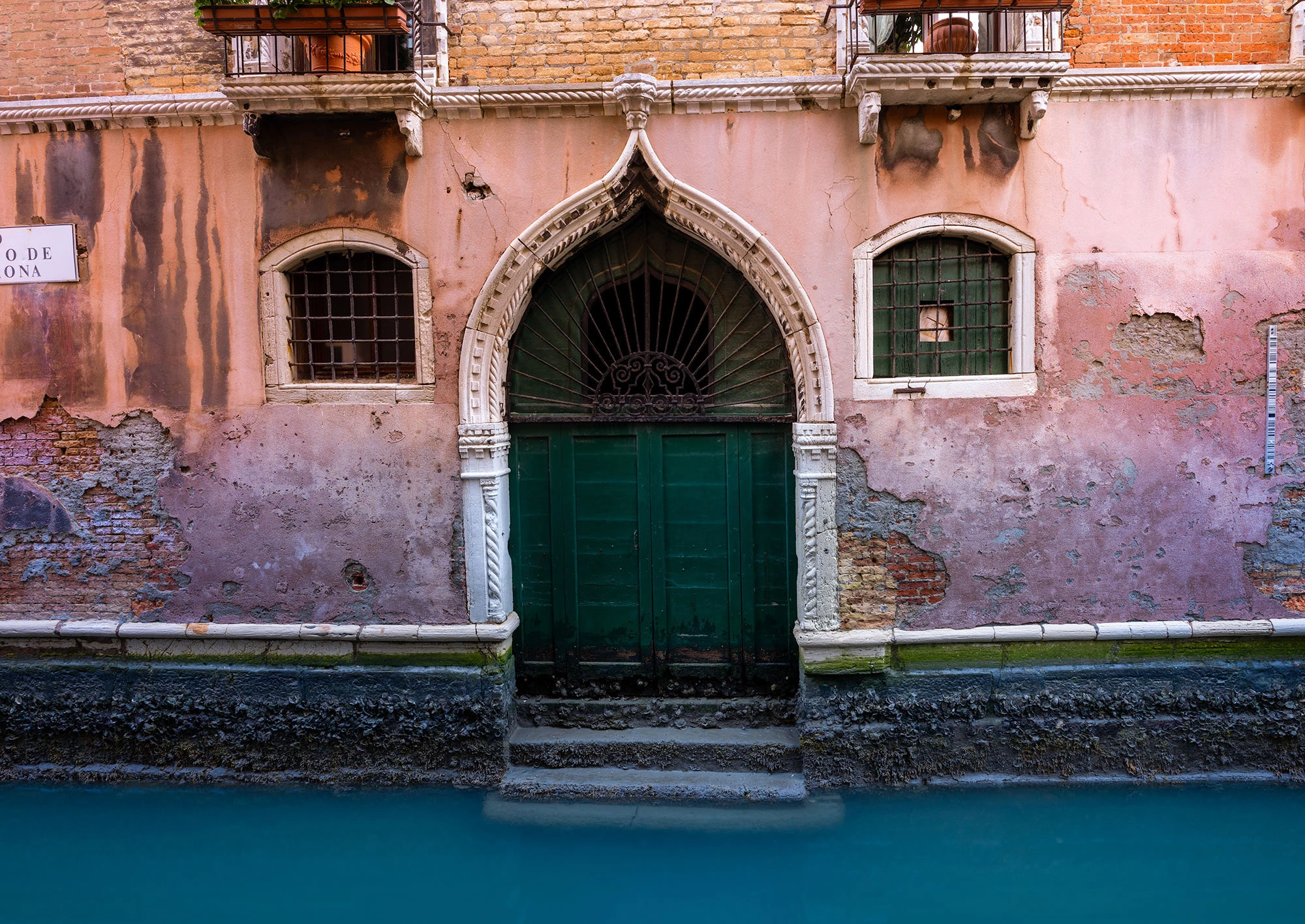 An old, weathered wooden door opens directly onto a calm Venice canal, framed by an aged brick building with small shuttered windows above.