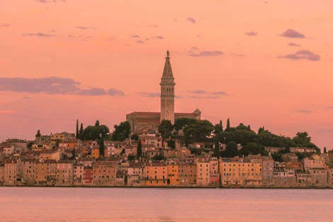 A predawn photo of the Rovinj cityscape in Croatia features the town's iconic silhouette with its bell tower rising prominently, bathed in the soft, bluish light of early morning, reflecting serenely off the calm Adriatic Sea.