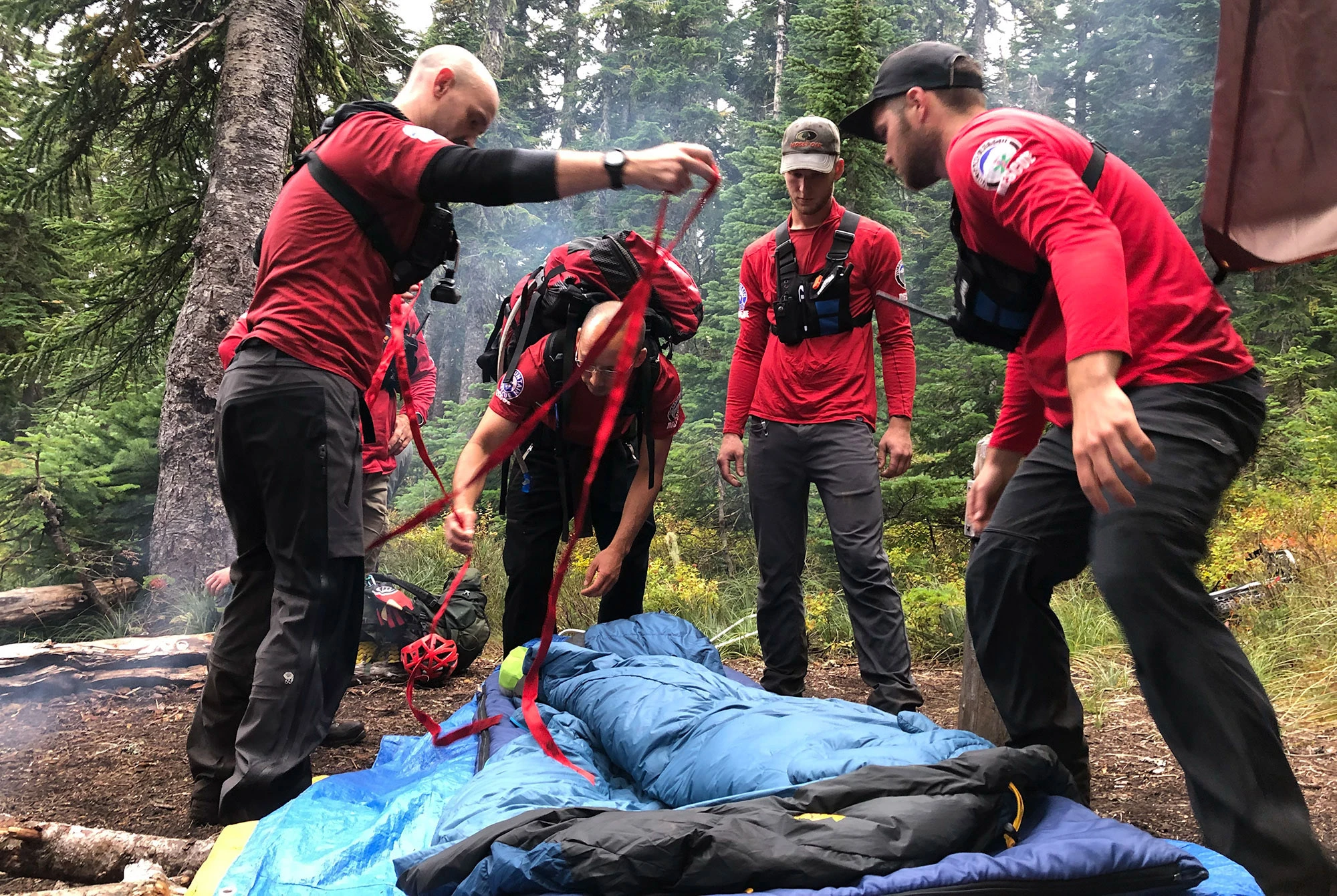 Members of the Volcano Rescue Team in red shirts and harnesses prepare a stretcher for evacuation after responding to a backpacking accident in the Indian Heaven Wilderness, Gifford Pinchot National Forest, Washington. One rescuer arranges red straps over a blue sleeping bag where the patient lies, while others stand by with gear ready, the alpine forest setting underscoring both the remoteness of the scene and the professionalism of the mountain rescue effort.